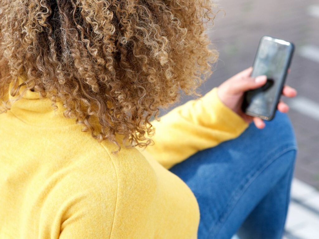 a woman with curly hair wearing a yellow sweater and blue jeans is seen from behind holding a mobile phone. She is sitting down.